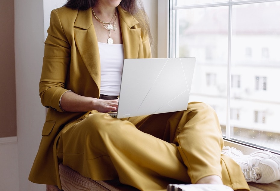 Une femme portant un costume jaune, assise près de la fenêtre, utilisant un Zenbook 14 OLED foggy silver