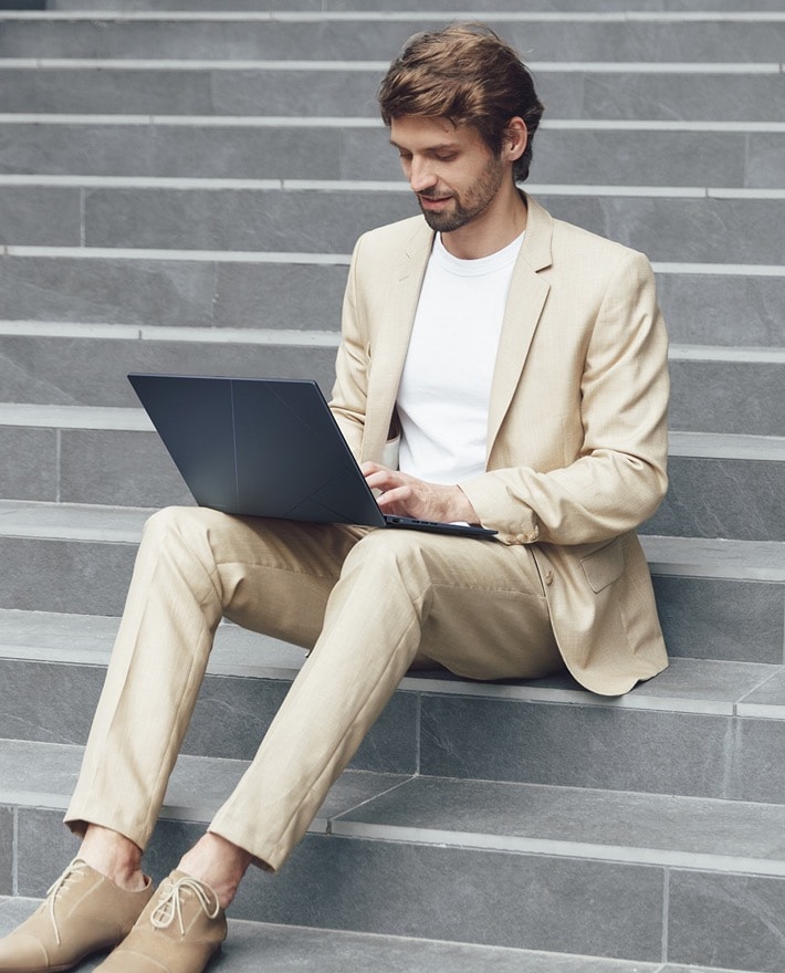 Un homme portant un costume beige assis sur les escaliers utilisant un Zenbook 14 OLED ponder blue